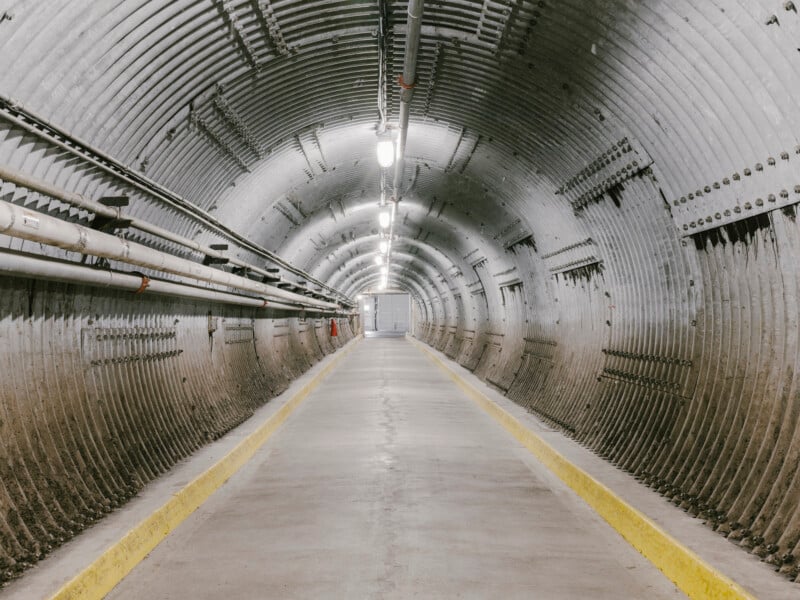 A long, empty concrete tunnel with corrugated metal walls, overhead lights, metal pipes along the sides, and yellow safety lines on both edges of the walkway, leading to a closed door at the end.