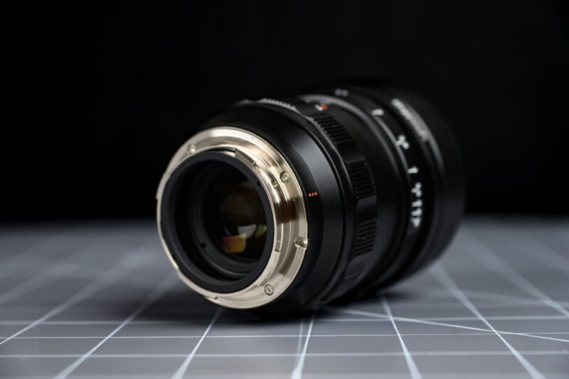 A close-up view of a camera lens placed on its side on a gray, grid-patterned surface, with the lens mount and glass elements clearly visible against a dark background.