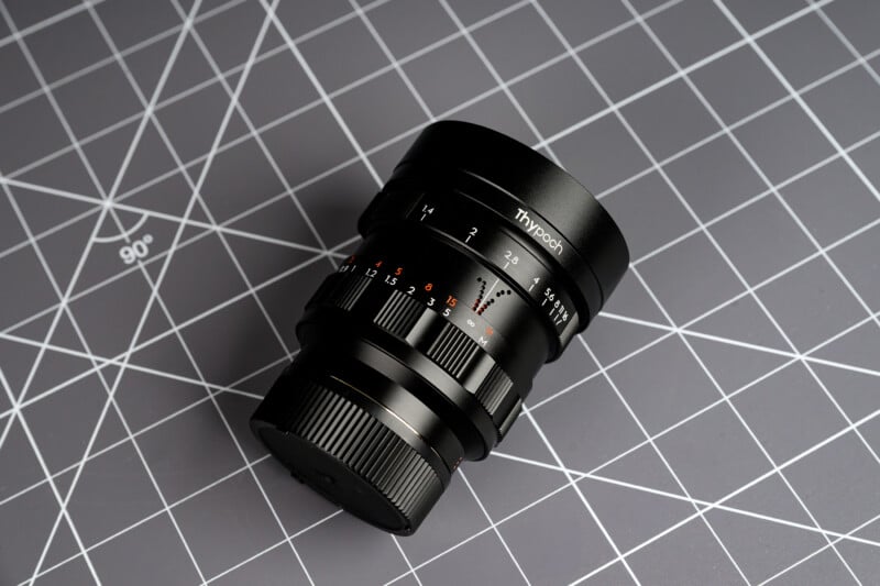 A black Thypoch camera lens with focus and aperture markings is placed on a gray gridded surface with white intersecting lines and angles.