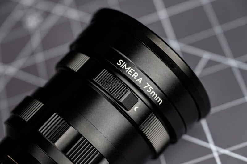 Close-up of a black camera lens labeled "SIMERA 75mm," with focus and aperture adjustment rings, placed on a surface featuring a white grid pattern.