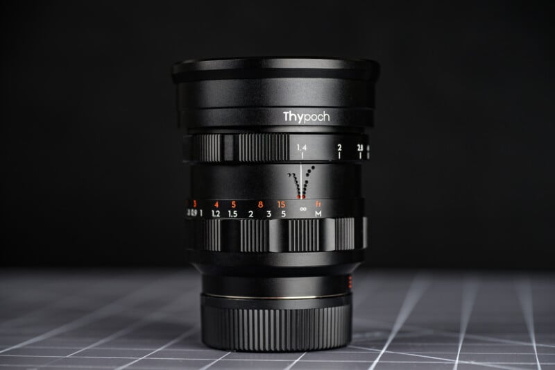 A black Thypoch camera lens stands upright on a grid-patterned surface with a dark background. The lens features various focus and aperture markings in white and orange.