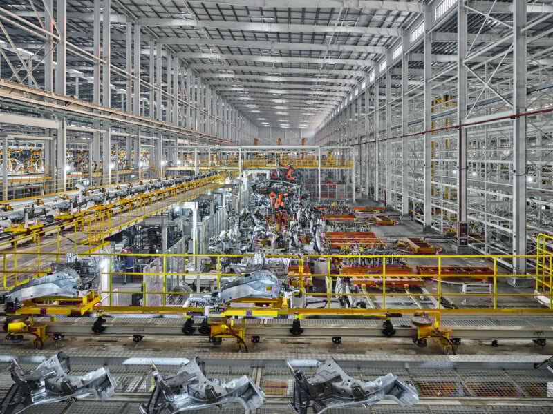 Large, modern automotive factory interior with high ceilings, machinery, robotic arms, metal frameworks, and vehicle parts being assembled on conveyor belts. The facility appears bright, organized, and industrial.