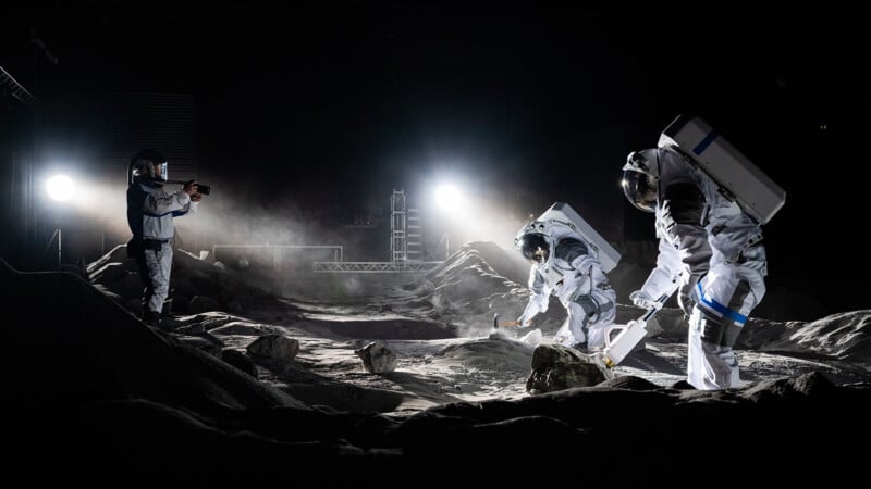 Three astronauts in white space suits are exploring a rocky, moon-like surface at night, with two collecting samples and one photographing them under bright artificial lights.