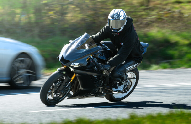 A person wearing a black outfit and helmet rides a black sport motorcycle at high speed on a road, with a blurred white car and greenery in the background, indicating motion.