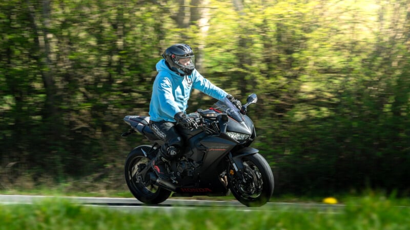 A person wearing a blue hoodie and helmet rides a black Honda motorcycle on a road with green trees and sunlight in the background.