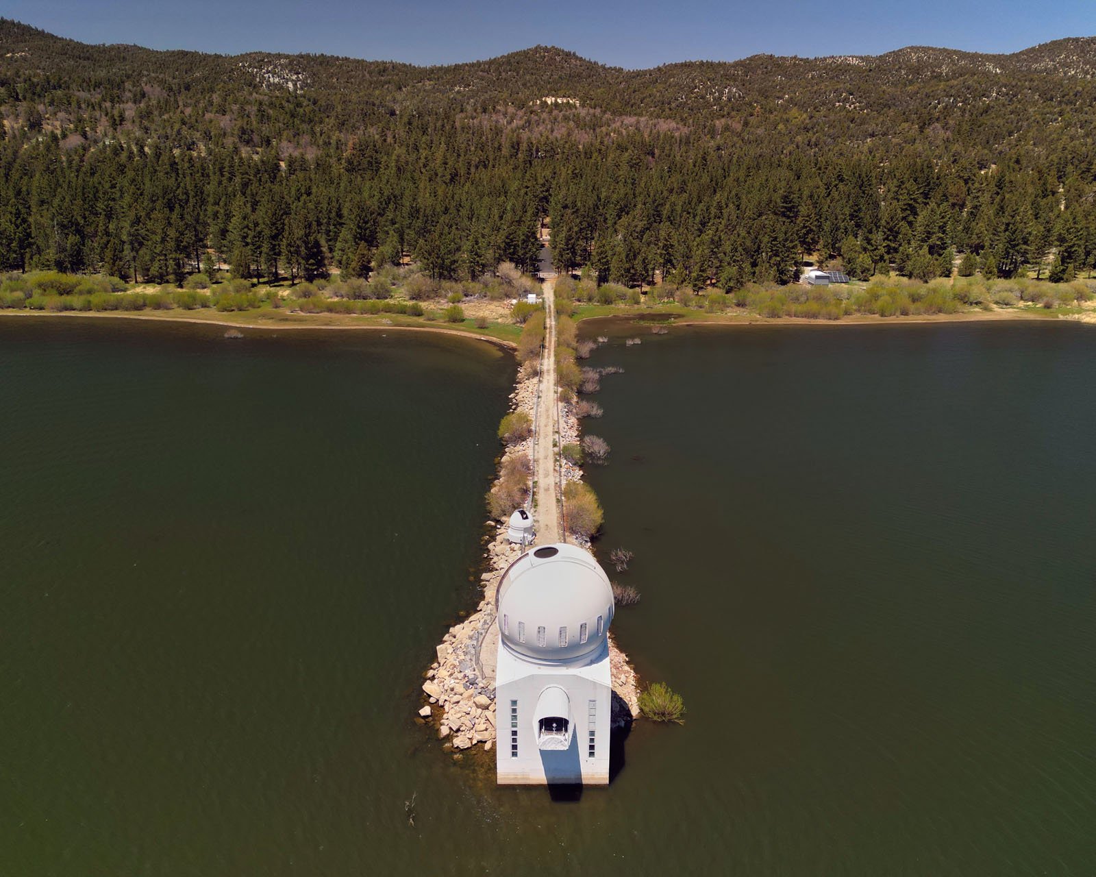 A domed building sits at the end of a narrow, tree-lined pathway extending into a lake, surrounded by forested hills under a clear sky.