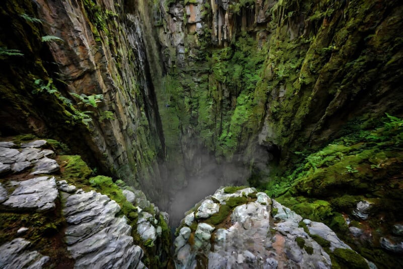 A deep, steep-sided sinkhole with moss and greenery covering the rocky walls, surrounded by rugged stone edges, and mist rising from the bottom.