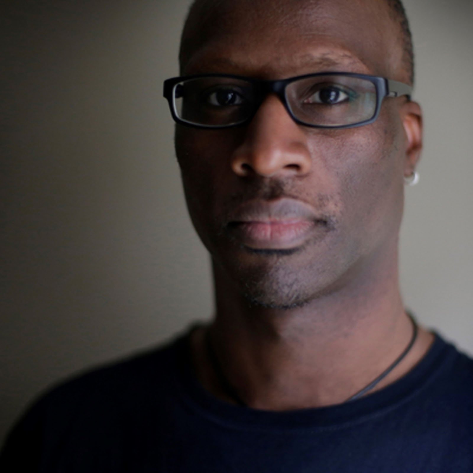 A person wearing black glasses and a dark shirt looks directly at the camera, with soft lighting highlighting one side of their face against a neutral background.