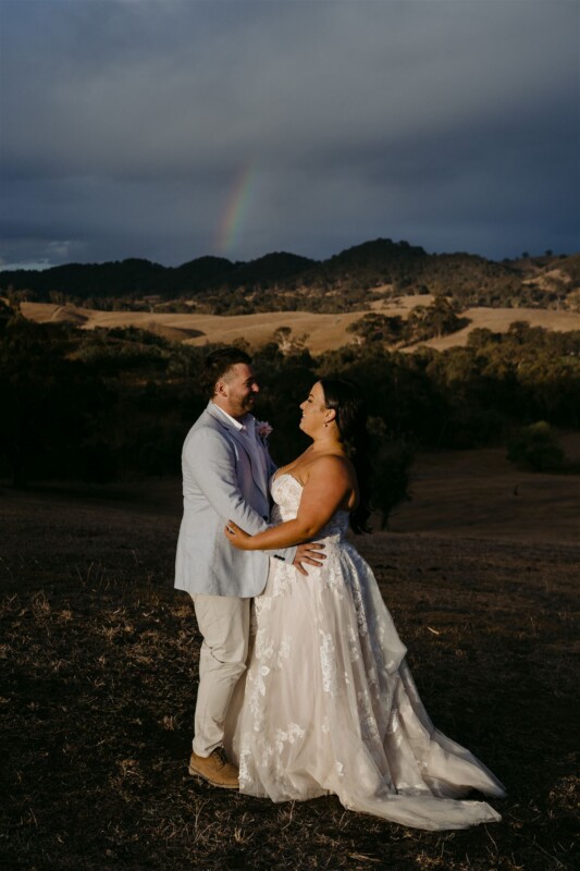 A couple in wedding attire stands smiling at each other on a grassy hillside at sunset, with dark hills and a faint rainbow in the cloudy sky behind them.