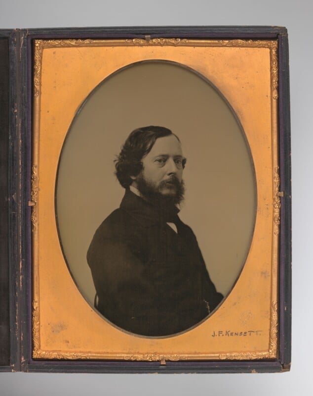 A black-and-white vintage portrait of a man with dark hair and a full beard, facing left. He wears a dark coat and is set in an ornate gold oval frame with a visible signature, "J.F. Kensett," at the bottom.