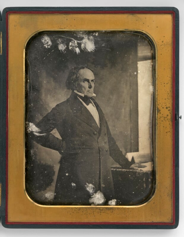 A sepia-toned daguerreotype of an older man in formal 19th-century clothing, standing with one hand inside his coat and looking off-camera. The photo is framed with a brass mat and shows signs of age and damage.