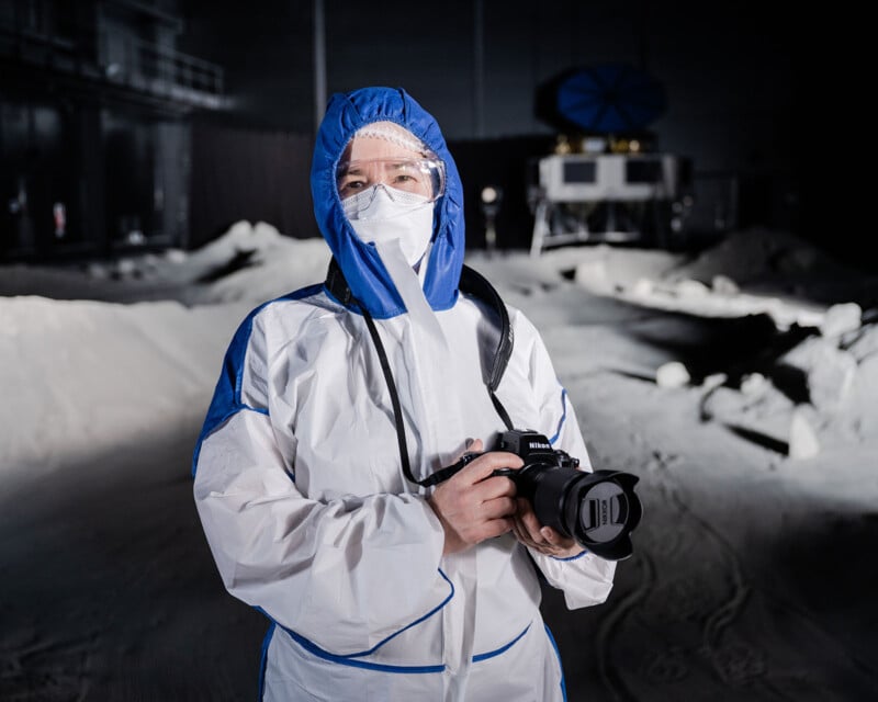 A person wearing a white and blue protective suit, face mask, and goggles holds a camera in a dimly lit, indoor environment resembling a lunar or space setting with rocks and equipment in the background.