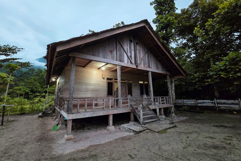 A rustic wooden house with a covered porch, raised on stilts, stands surrounded by lush green trees and plants. The lights on the porch are on, and the area around the house appears sandy and natural.