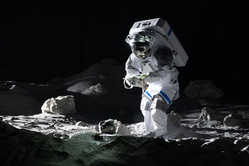 An astronaut in a white spacesuit collects rock samples on a rocky, gray lunar surface under dramatic lighting, with large boulders and shadows surrounding them.