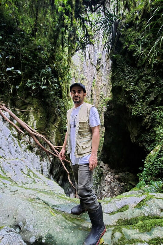 A man wearing a beige vest, white shirt, and rubber boots stands on mossy rocks in a lush, green forested ravine surrounded by tall plants and vines.