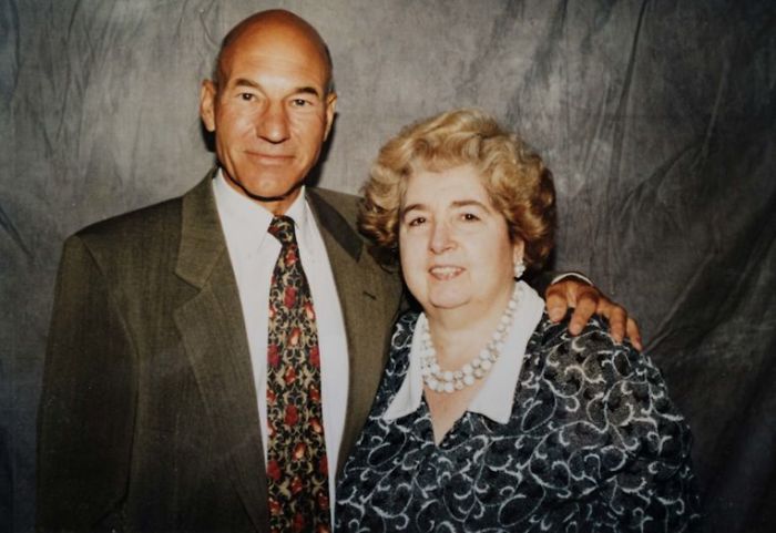 A man in a suit and patterned tie stands beside a woman with short, curly hair wearing a patterned blouse and pearl necklace. They both smile at the camera against a gray backdrop.