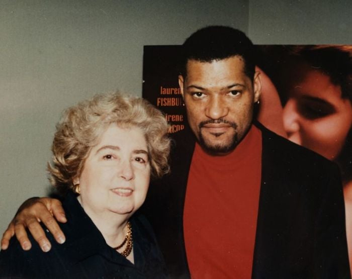 A man in a red shirt with his arm around an older woman with light hair, both smiling at the camera. There is a partially visible poster in the background with indistinct text and images.