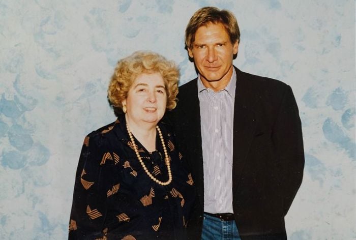 A woman with curly blonde hair and a patterned dress stands next to a man in a dark blazer and striped shirt, both smiling in front of a light blue, softly patterned background.