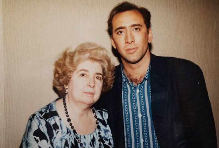 A man in a patterned shirt and dark jacket stands next to an elderly woman with short, curly hair and a floral blouse. They pose together against a plain, beige background.