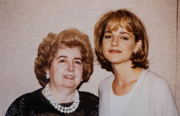 Two women stand side by side, smiling at the camera. The older woman has curly light hair and wears a black top with pearl jewelry, while the younger woman has short blonde hair and a light-colored sweater. Plain background.