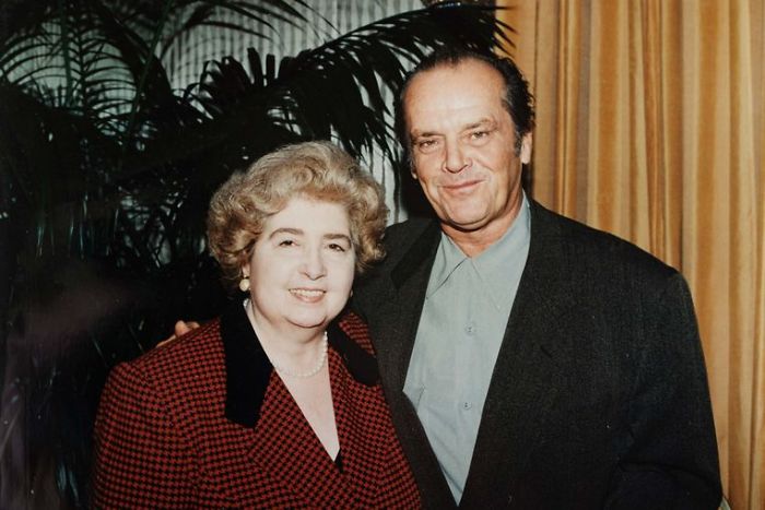 A smiling older woman with curly hair stands next to a smiling man with short hair and a dark jacket. They are indoors, with a curtain and large green plant in the background.