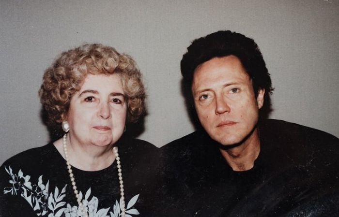 An older woman with curly gray hair and pearl jewelry sits next to a middle-aged man with dark hair. Both are looking at the camera with neutral expressions, against a plain background.