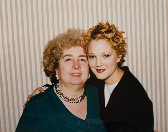 Two women pose closely together, smiling at the camera. One has short curly hair and wears a teal top with statement jewelry; the other has short, styled blonde hair and wears a dark jacket. The background features vertical striped wallpaper.