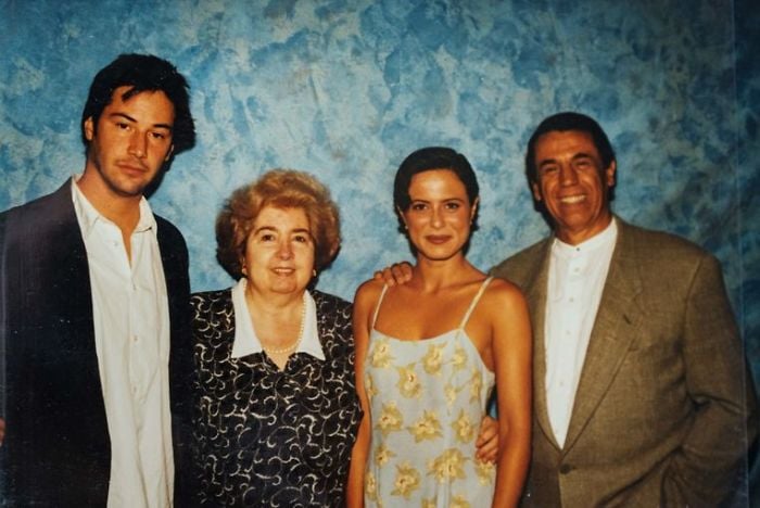 Four people stand closely together, posing and smiling in front of a blue textured backdrop. Two men wear suits, while the two women are dressed in a patterned blouse and a floral dress.