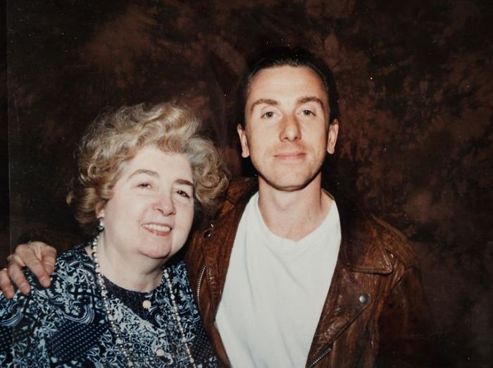 A smiling older woman with curly hair stands next to a younger man in a brown leather jacket and white t-shirt, posing in front of a brown, textured backdrop. The man has his arm around the woman's shoulder.