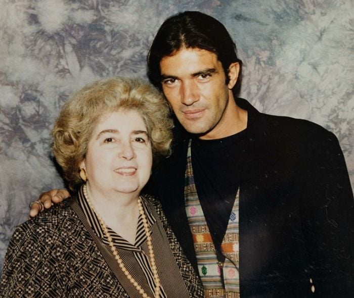 A smiling older woman with curly blond hair stands next to a younger man with dark hair, wearing a patterned vest and black jacket, in front of a gray marbled backdrop.