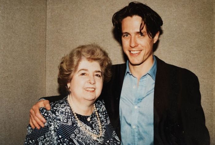 A smiling older woman with short, curly hair and a patterned dress stands beside a younger man in a blue shirt and black blazer, posing together in front of a plain wall. The man has his arm around the woman's shoulders.