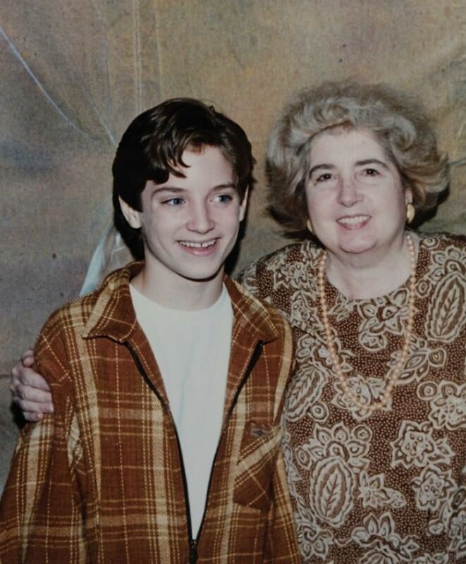 A smiling teenage boy in a brown plaid jacket stands next to an older woman with gray hair wearing a patterned brown dress. They both face the camera and smile, with the woman’s arm around the boy’s shoulder.