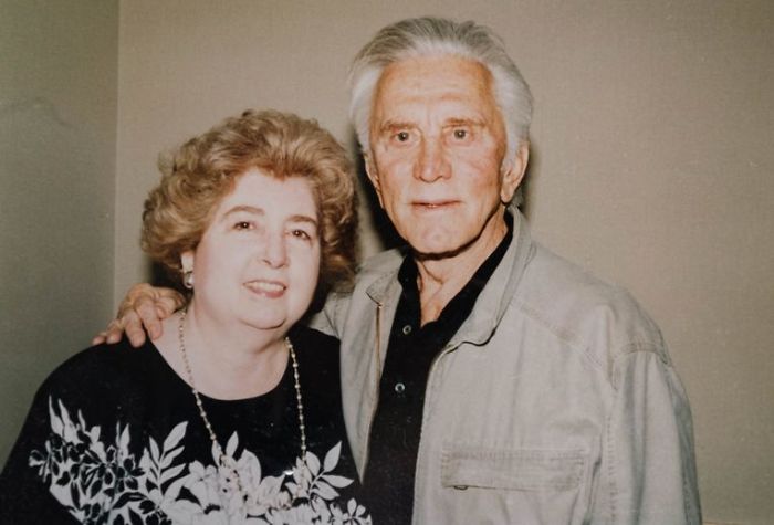 An older man with gray hair and a light jacket stands next to a woman with curly hair and a floral-patterned top. They are smiling and posing together against a plain beige background.