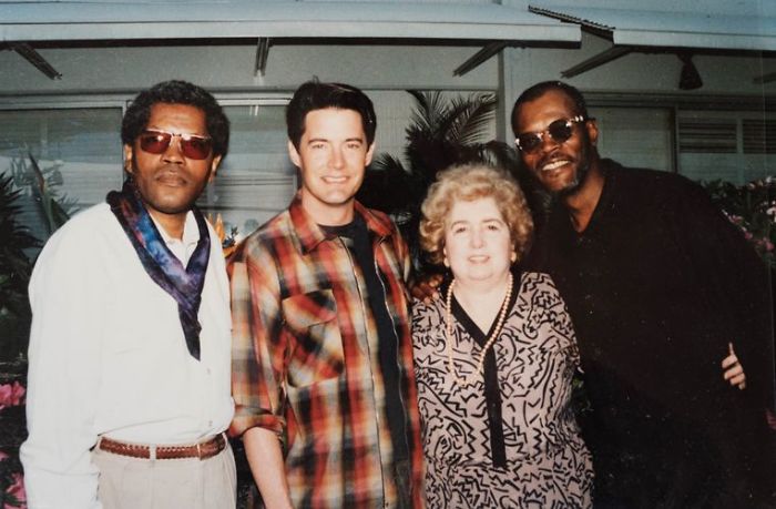 Four adults, three men and one woman, stand closely together smiling outdoors. The men wear sunglasses and casual shirts; the woman wears a patterned dress. There are plants and a building with large windows in the background.