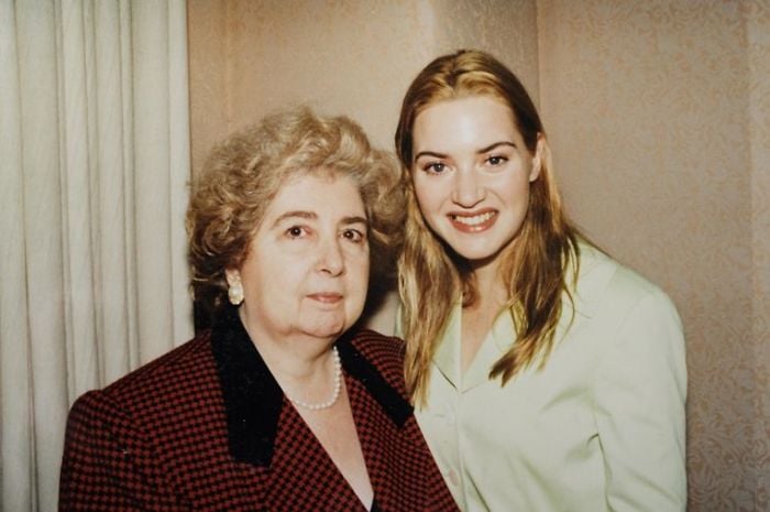 Two women pose together indoors. The older woman has short, curly gray hair and wears a red checkered jacket with a pearl necklace. The younger woman has long blonde hair and wears a light green top, smiling at the camera.