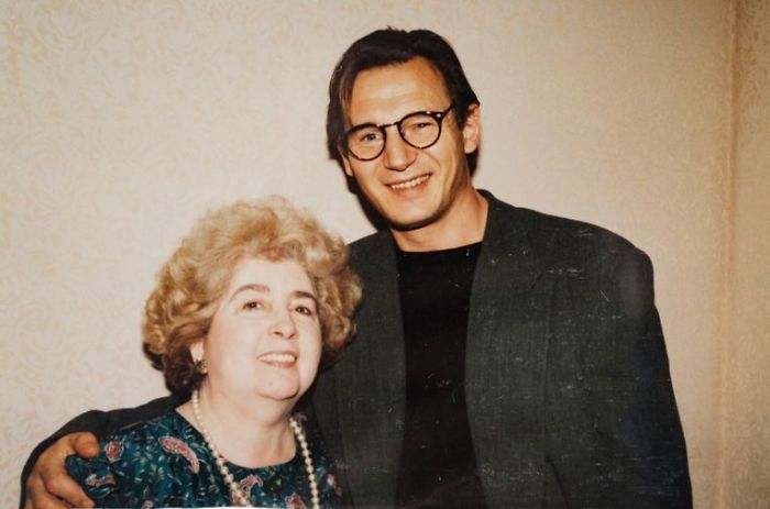 A smiling man with glasses and a dark jacket stands next to an older woman with curly hair and a floral top. They both look at the camera, and he has his arm around her shoulders. The background is a light patterned wall.