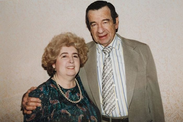 An older man in a suit and tie stands with his arm around an older woman in a patterned dress and pearl necklace; both are smiling in front of a light-colored patterned wall.