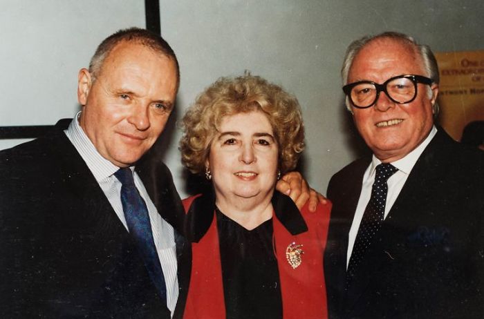 Three older adults in formal attire pose together, smiling for the camera. The man on the left is bald, wearing a suit and tie; the woman in the middle has curly hair and wears a red jacket; the man on the right wears glasses and a suit.
