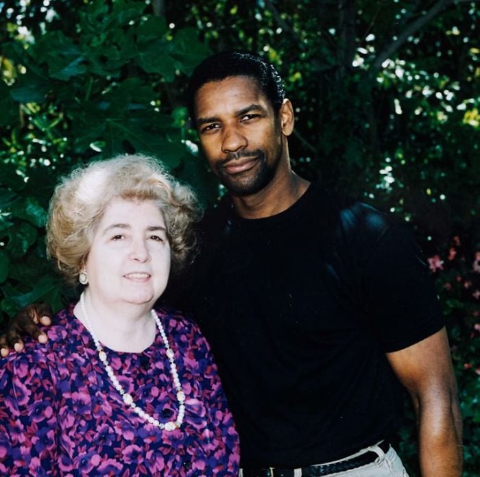 A man in a black t-shirt stands outdoors with his arm around an older woman in a purple patterned blouse and pearl necklace. They are both smiling, with greenery and trees in the background.