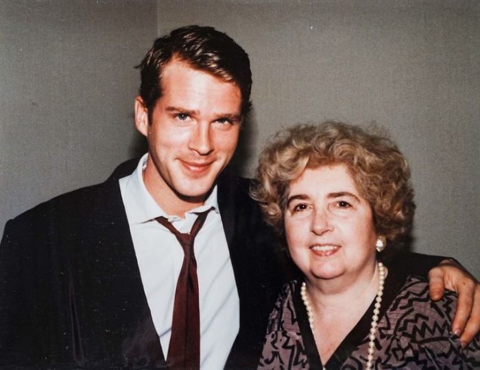 A young man in a suit and tie stands with his arm around an older woman with curly gray hair, wearing a pearl necklace and patterned top. Both are smiling and facing the camera against a plain background.