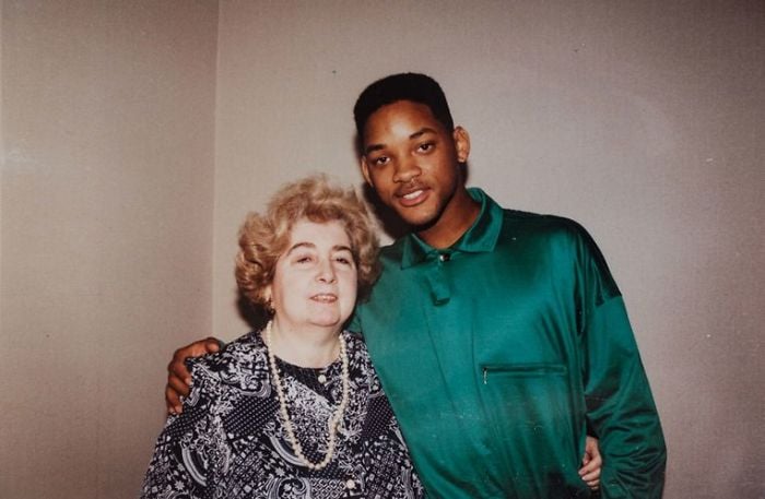 A young man in a green shirt stands with his arm around an older woman with curly, light hair and a patterned dress, both smiling and posing against a plain background.