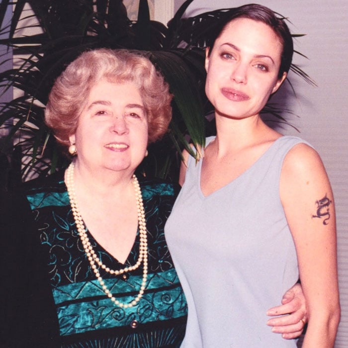 Two women standing close together and smiling. The older woman has gray hair, is wearing a patterned dress with pearl necklaces. The younger woman has dark hair in a bun, is wearing a sleeveless top, and has a tattoo on her upper arm.