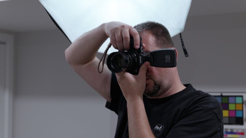 A person in a black shirt holds a camera up to their face, preparing to take a photo in a studio with a large softbox light and a color calibration chart in the background.
