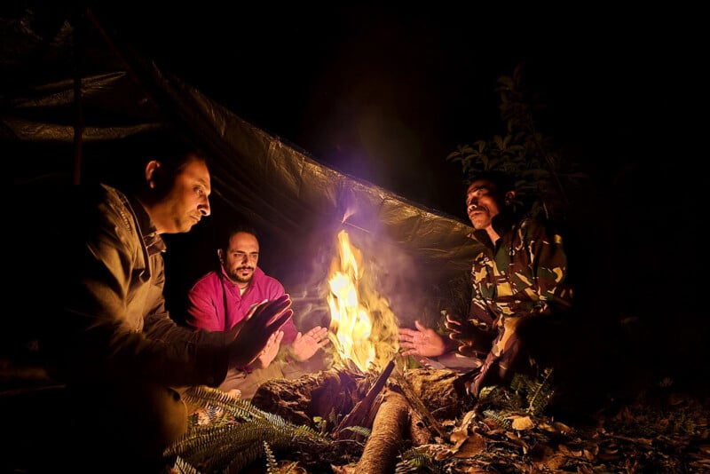 Three people sit around a campfire at night, warming their hands. A tarp or tent is set up behind them, and the surrounding area is dark except for the firelight illuminating their faces.