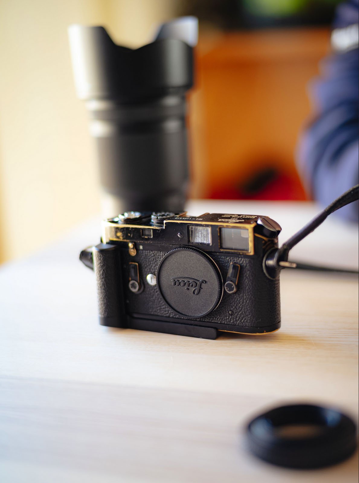 A black film camera with a lens cap labeled "Leica" rests on a light wooden surface. A blurred camera lens and a person's arm are visible in the background.