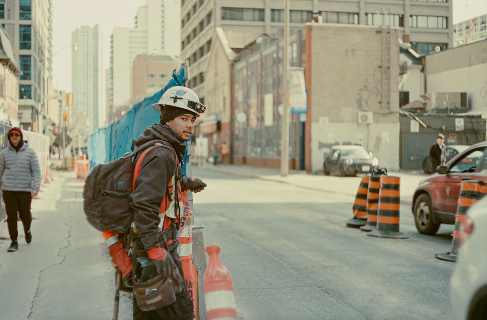 A construction worker in safety gear stands on a city street surrounded by traffic cones and barriers, with tall buildings in the background and pedestrians walking nearby.