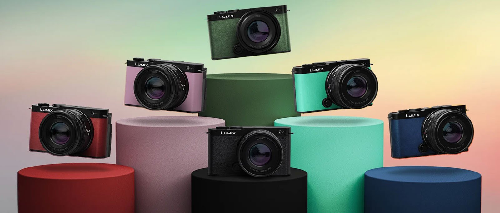 Seven Panasonic Lumix cameras in various colors are displayed on colorful cylindrical pedestals against a soft gradient background.