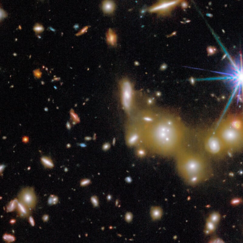 A field of distant galaxies and stars glows against the darkness of space, with a bright blue star and lens flare at the top right and several fuzzy, yellowish elliptical galaxies near the center.