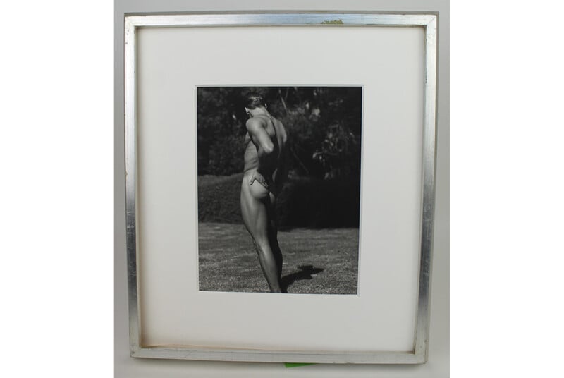 Black and white photo of a person in swimwear standing outdoors on grass, seen from the side with their head turned down, displayed in a white mat and silver frame.