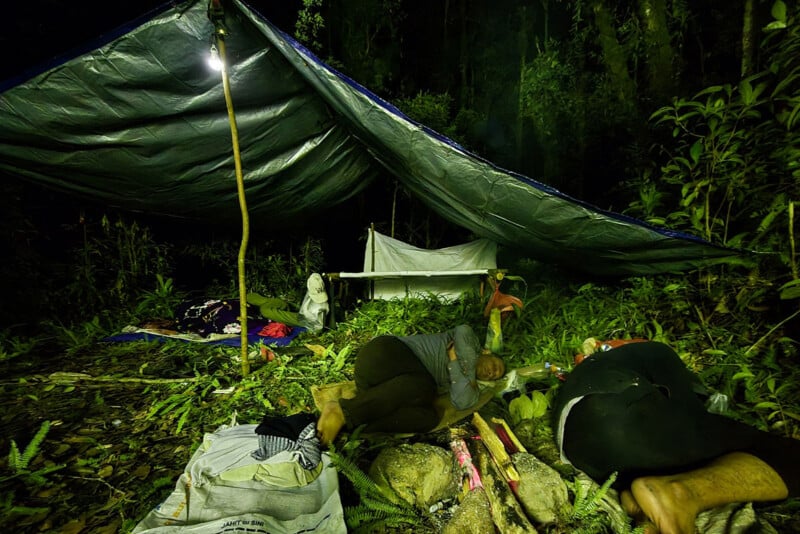 Two people sleep under a makeshift tarp shelter in a forest at night, illuminated by a hanging light; bags and belongings are scattered around on the green foliage and ground.